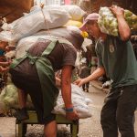 Trabalhadores carregam um carrinho com produtos em um movimentado mercado de rua.