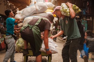 Trabalhadores carregam um carrinho com produtos em um movimentado mercado de rua.